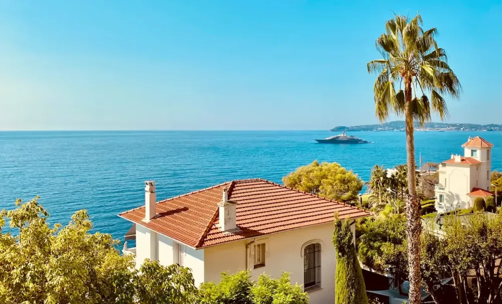 Villa méditerranéenne blanche au toit de terre cuite face à la mer bleue, entourée d'arbres.